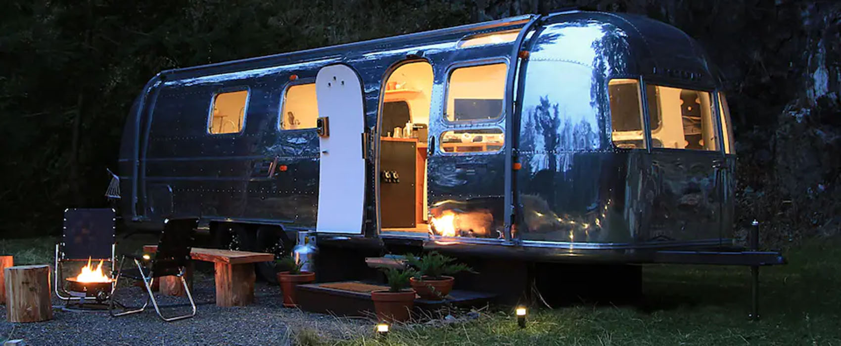 Long chrome airstream trailer parked behind a firepit seating area, with lights on inside at dusk.