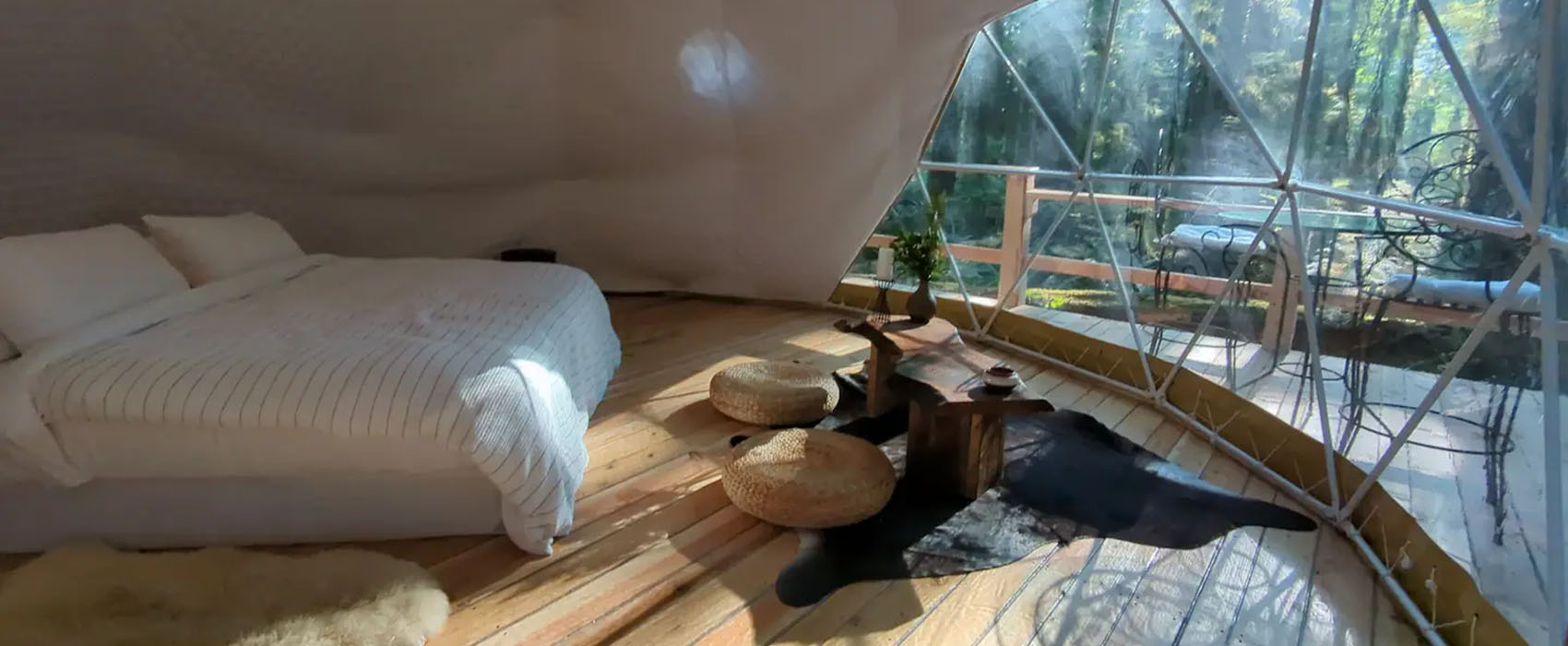 Windowed geodesic dome with wooden floors, large bed, fur rugs and seating area inside. View out the window shows a patio with seating area and forest view.